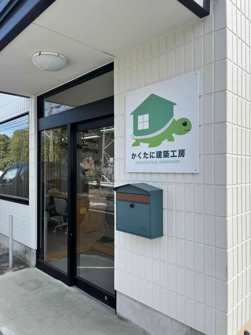 かくたに建築工房の入口の白い外壁に取り付けた、緑の亀と家のロゴが入ったプレート看板